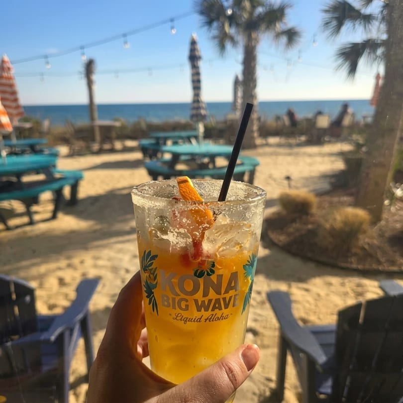 Hand holding a Kona Big Wave cocktail with a beach and ocean background.