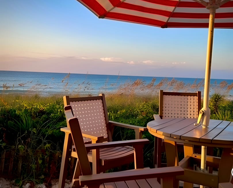 Outdoor dining area with chairs, table, and red-striped umbrella overlooking the ocean at sunset.