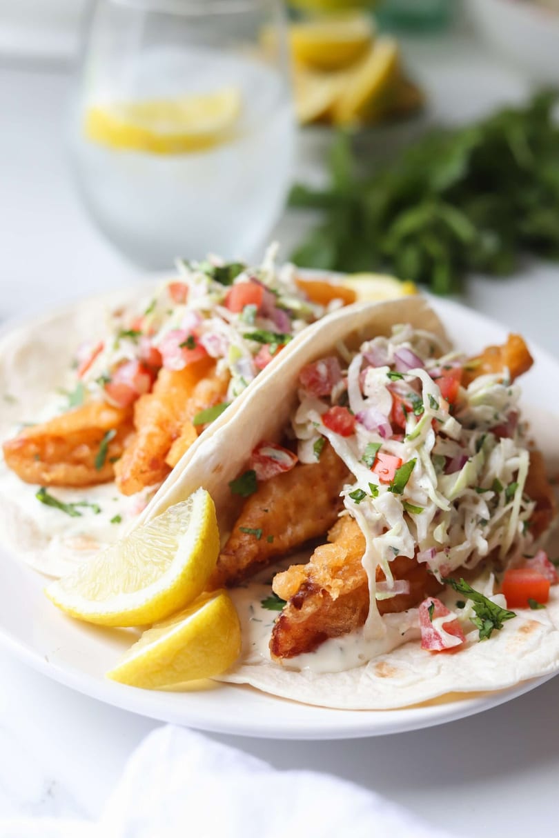 Two fish tacos with crispy fish, coleslaw, salsa, and lemon wedges on a white plate.