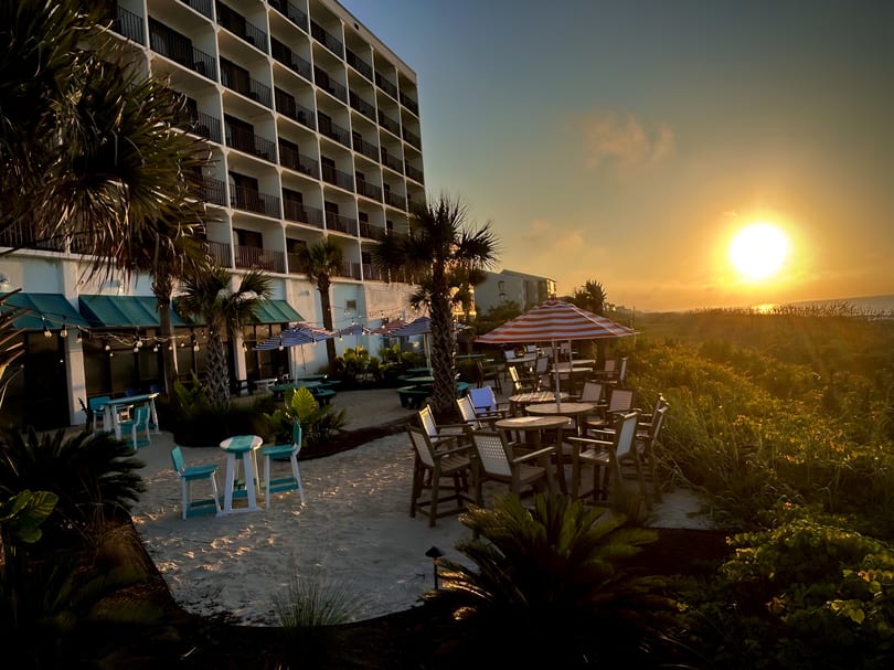 Sunset over a beachside restaurant patio with palm trees and hotel in the background.