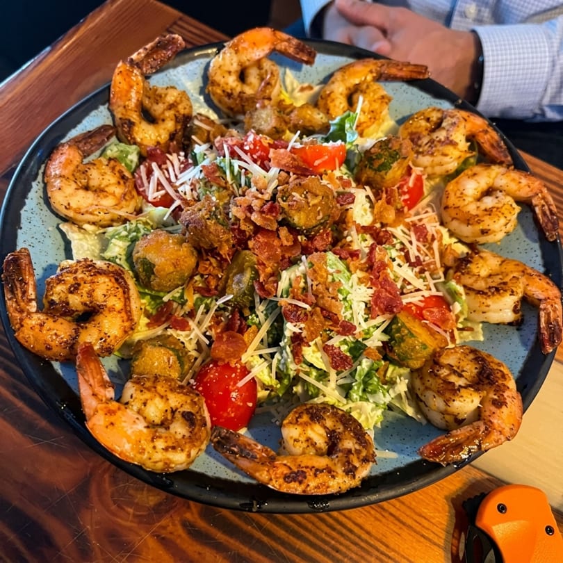 A large Caesar salad topped with grilled shrimp, bacon, cherry tomatoes, and fried Brussels sprouts.