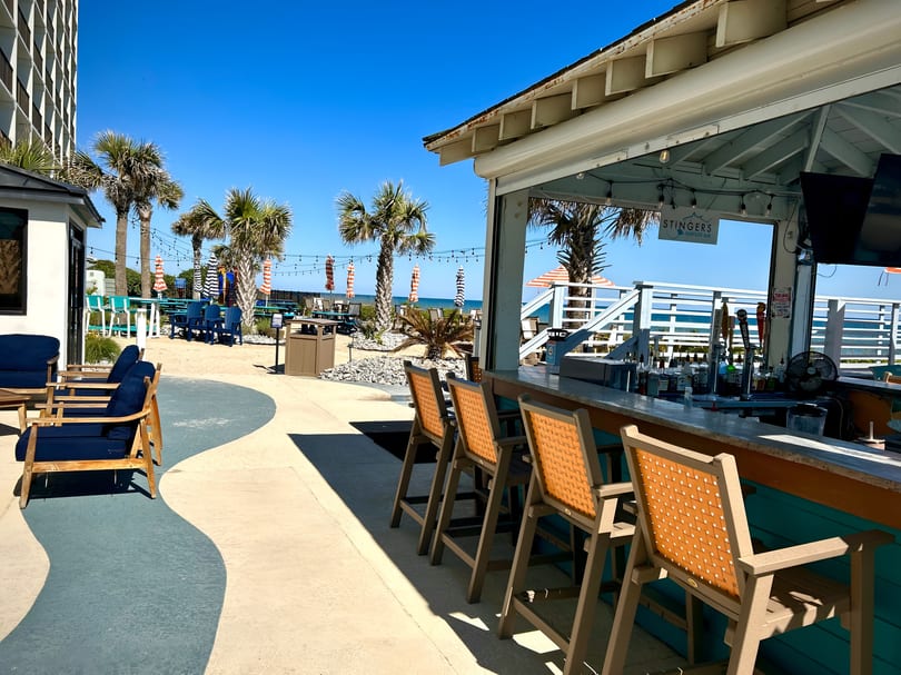 Outdoor bar with orange stools, palm trees, and ocean views under a clear blue sky.